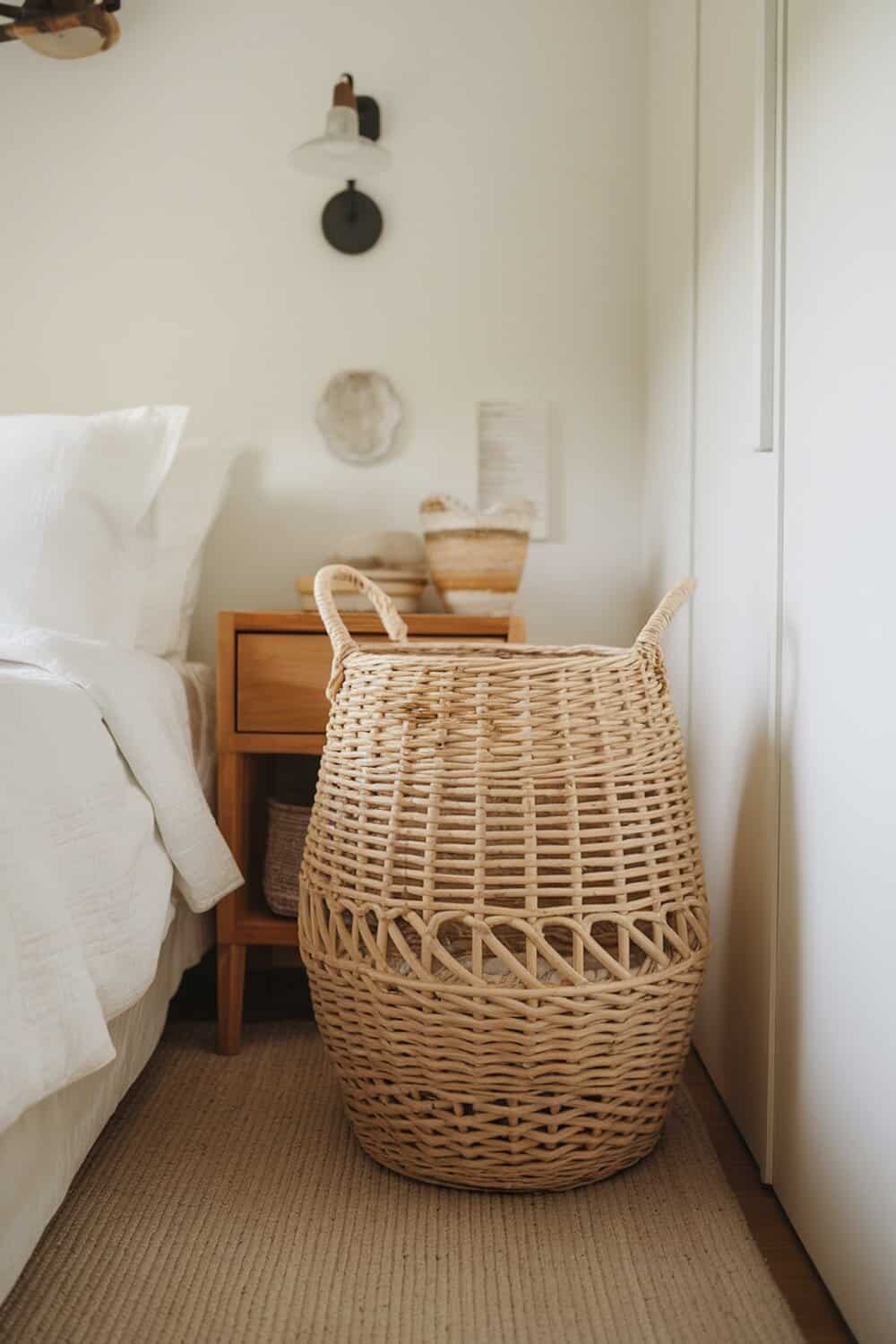 A woven laundry basket beside a bed in a cozy bedroom setting.