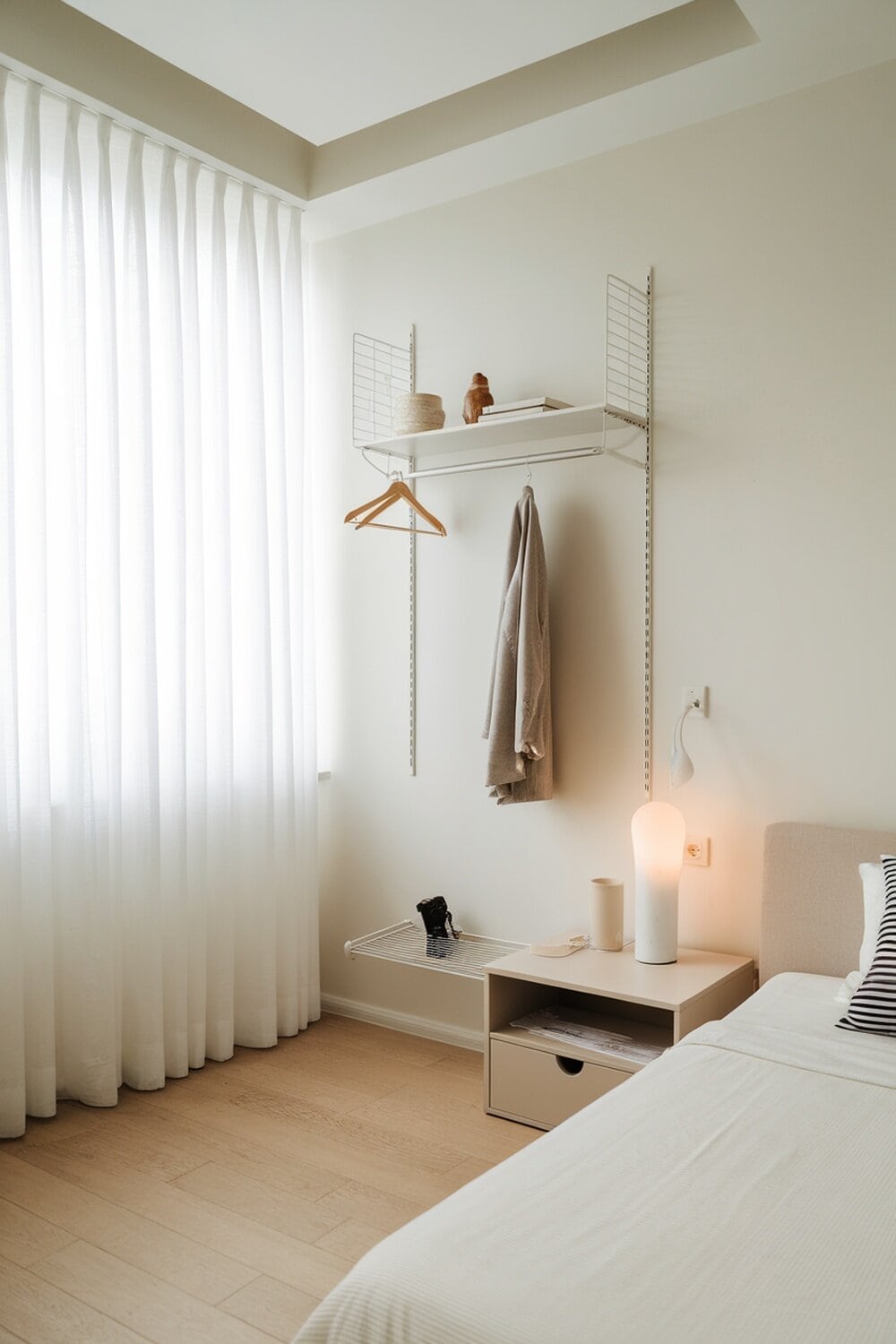 A minimalist bedroom with a wall shelf, a small nightstand, and natural light.