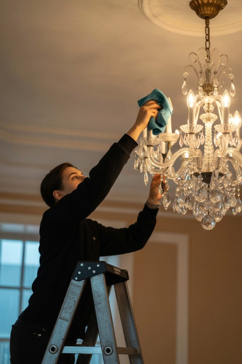 A person cleaning a chandelier while standing on a ladder.