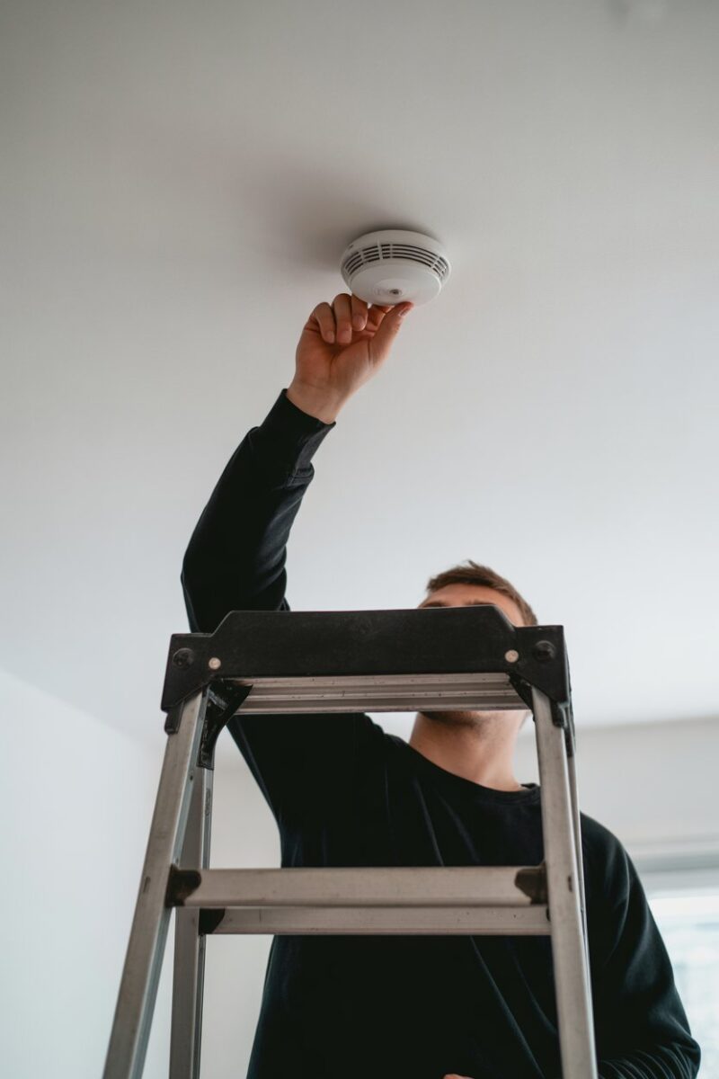 A person testing a smoke detector on the ceiling while standing on a ladder.