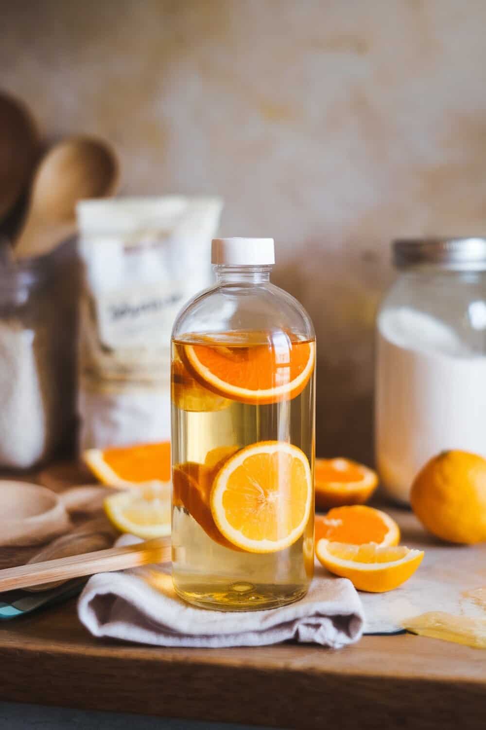 A bottle of homemade citrus cleaner with orange slices, surrounded by citrus fruits and cleaning supplies.