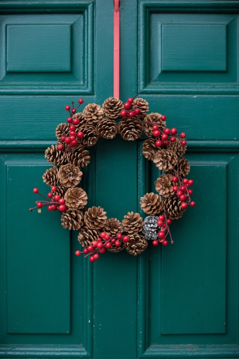 A wreath made of pinecones and red berries hanging on a teal door.