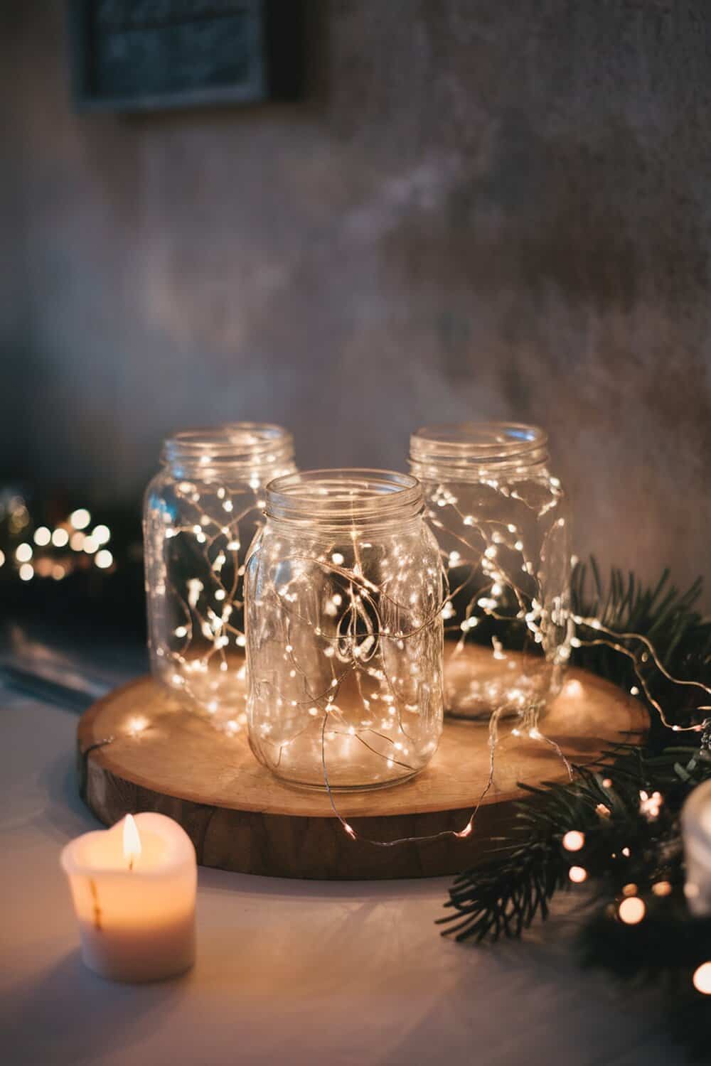 Three glass jars filled with fairy lights on a wooden surface, with a lit candle nearby.