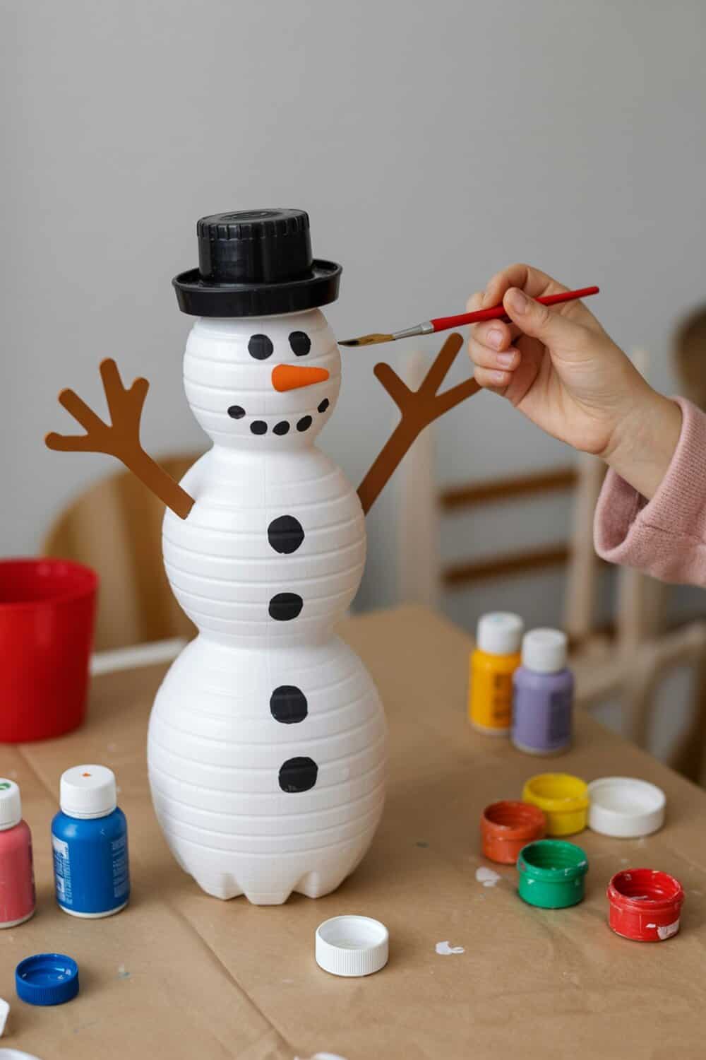 A child painting a snowman made from recycled bottles, with colorful paint and supplies around.