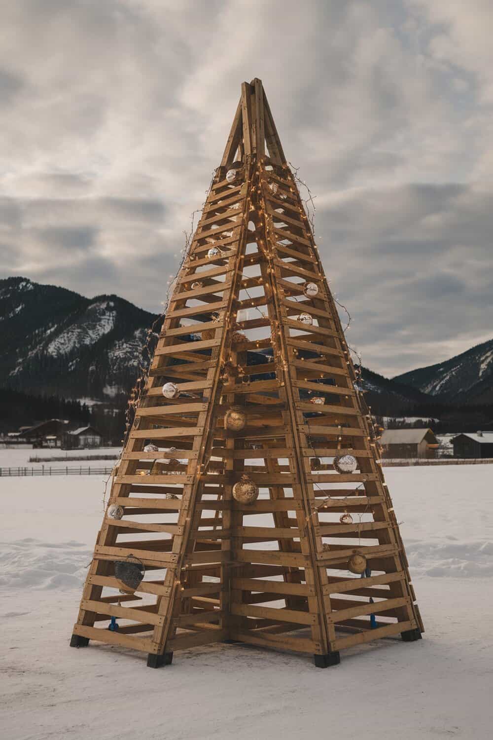 A Christmas tree made from wooden pallets, decorated with lights and ornaments, set against a snowy landscape.