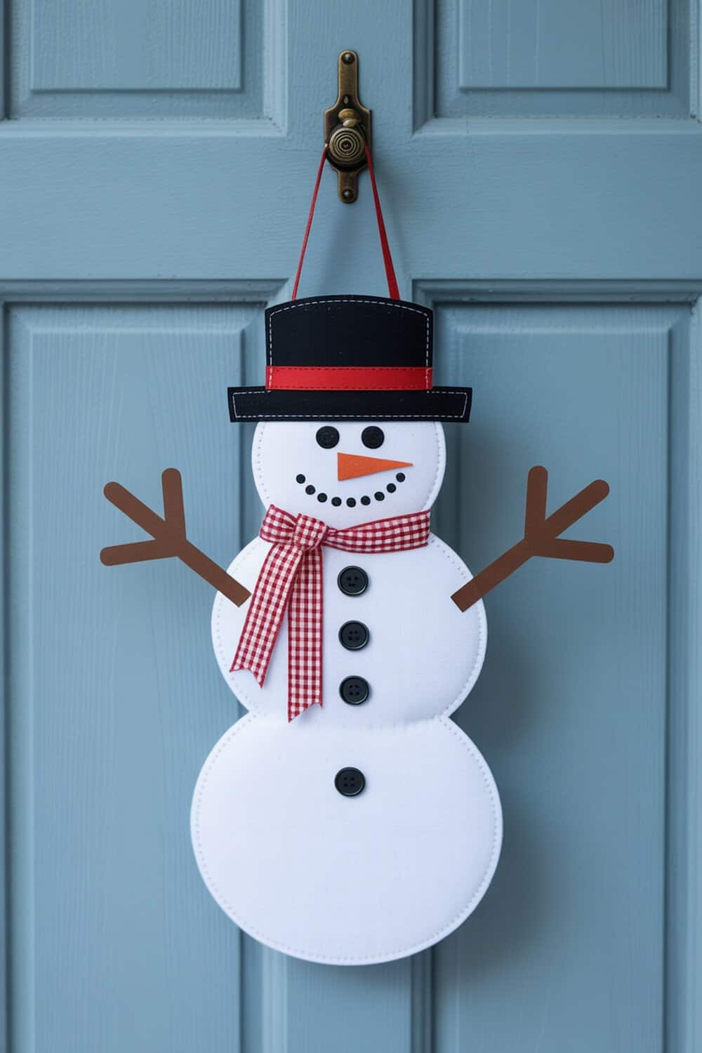 A snowman door hanger with a black hat, red scarf, and buttons, hanging on a blue door.