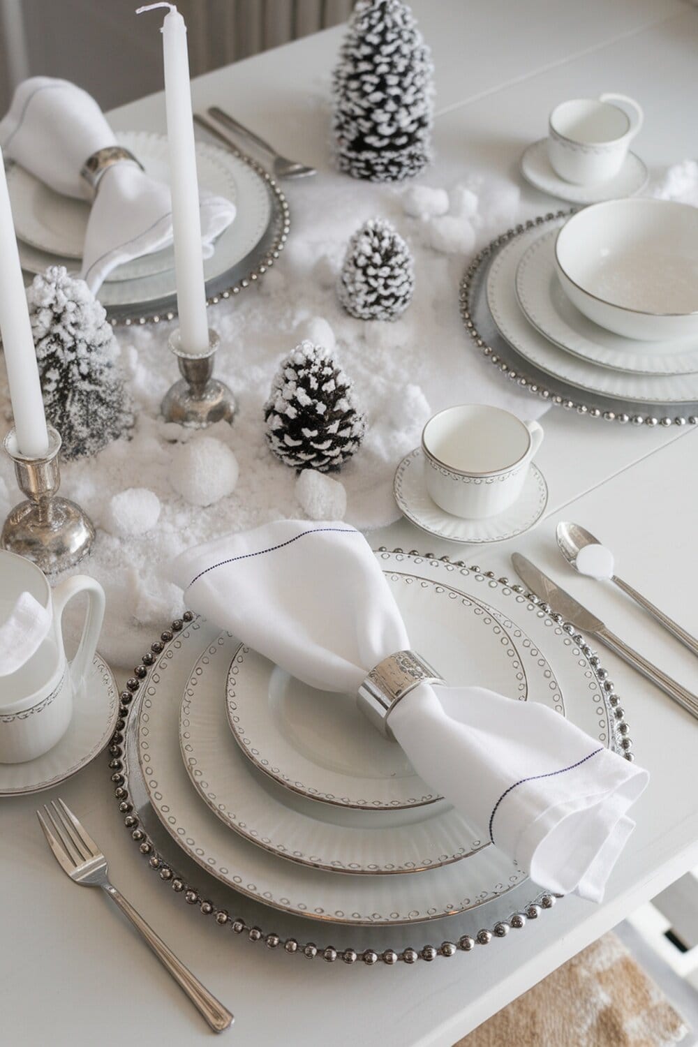 A snowy white table setting with elegant plates, silver accents, and decorative pine trees.