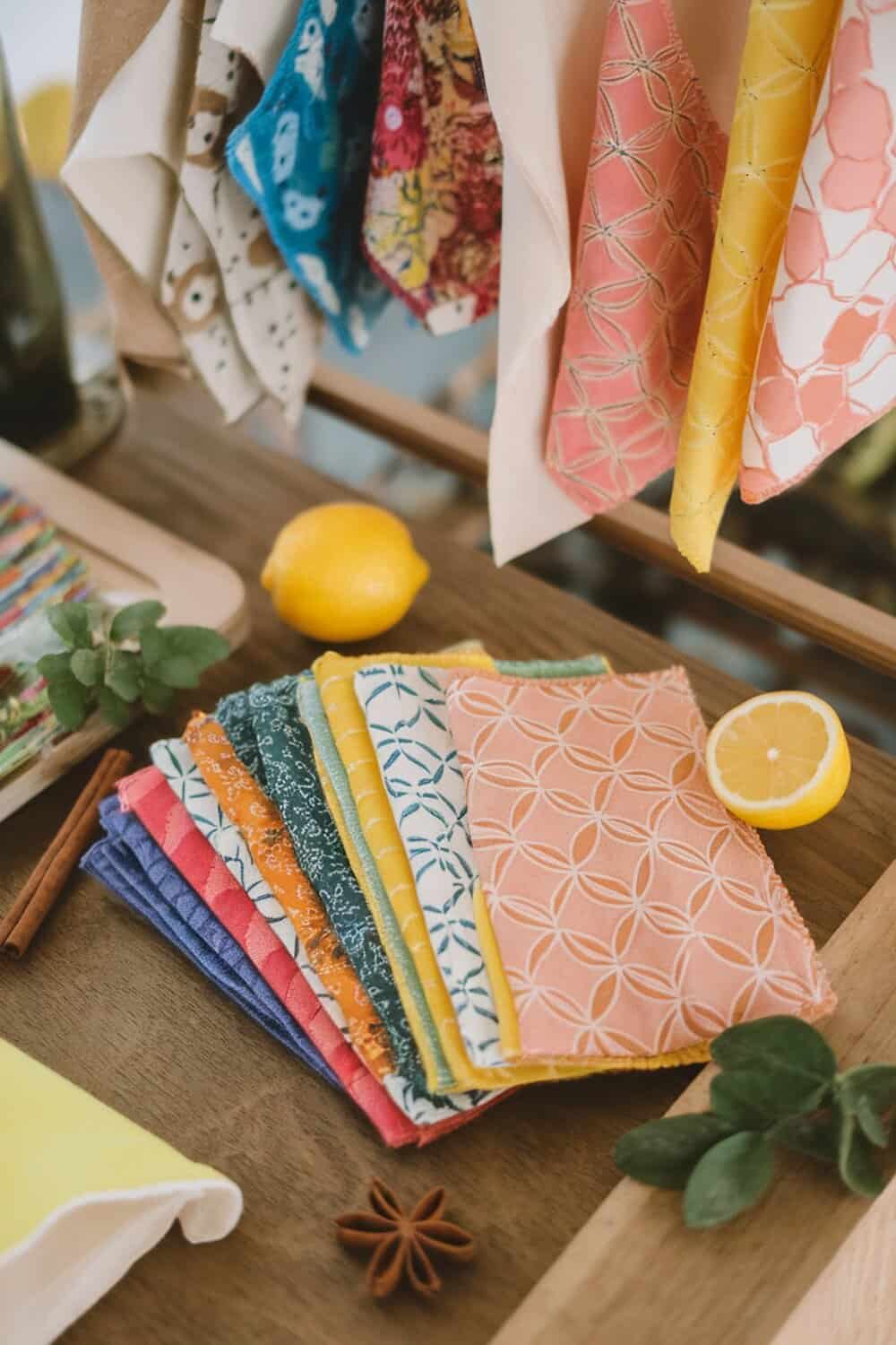 Colorful reusable beeswax wraps displayed on a wooden surface with a lemon and herbs.