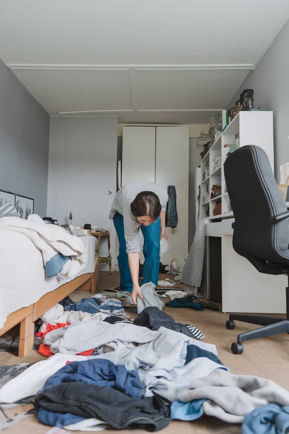A person tidying up a messy room by picking up clothes from the floor.