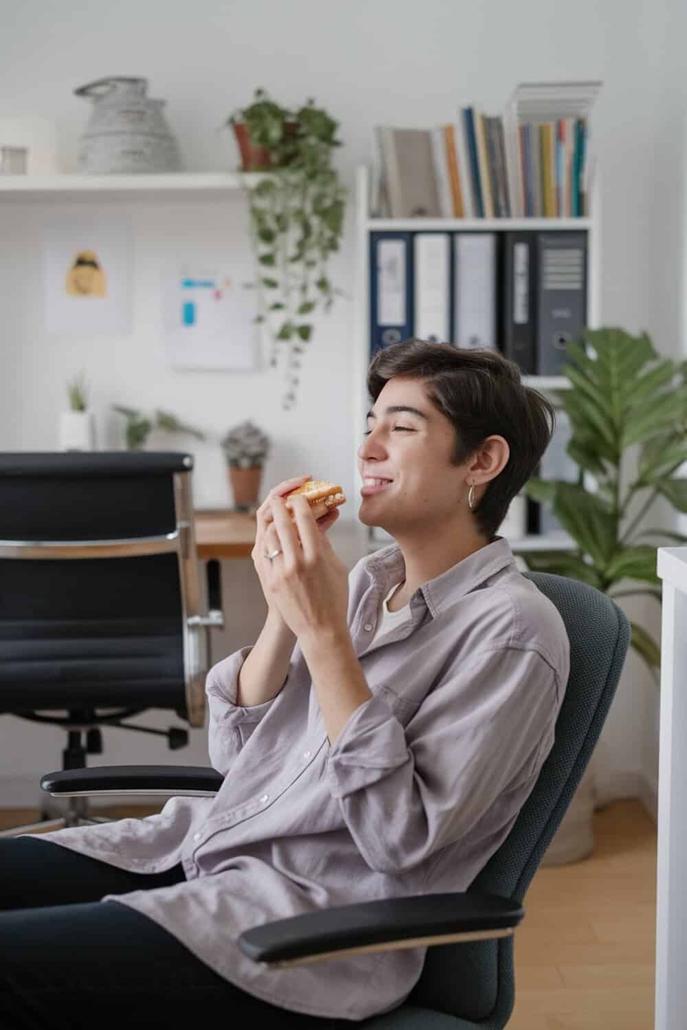 A person sitting in a chair, smiling and enjoying a snack in a tidy room.