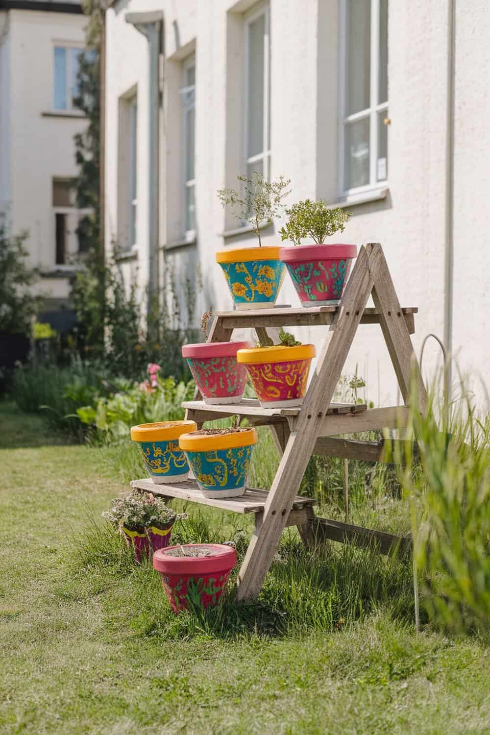 Colorful hand-painted plant pots displayed on a wooden ladder in a garden.