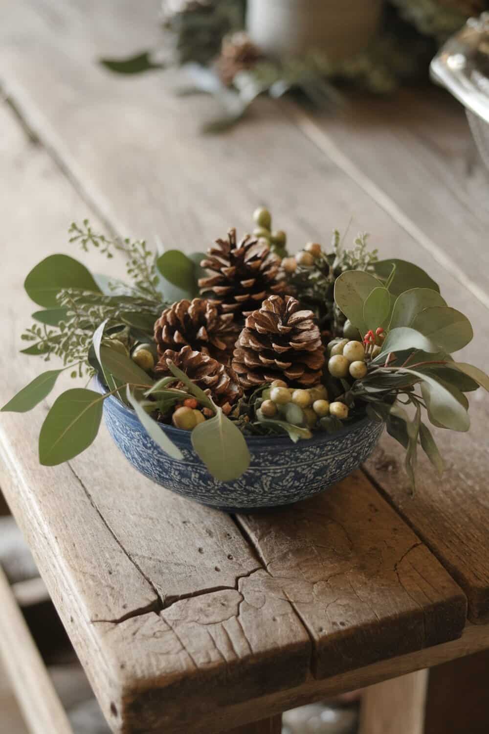A decorative bowl filled with pinecones and greenery on a wooden table.