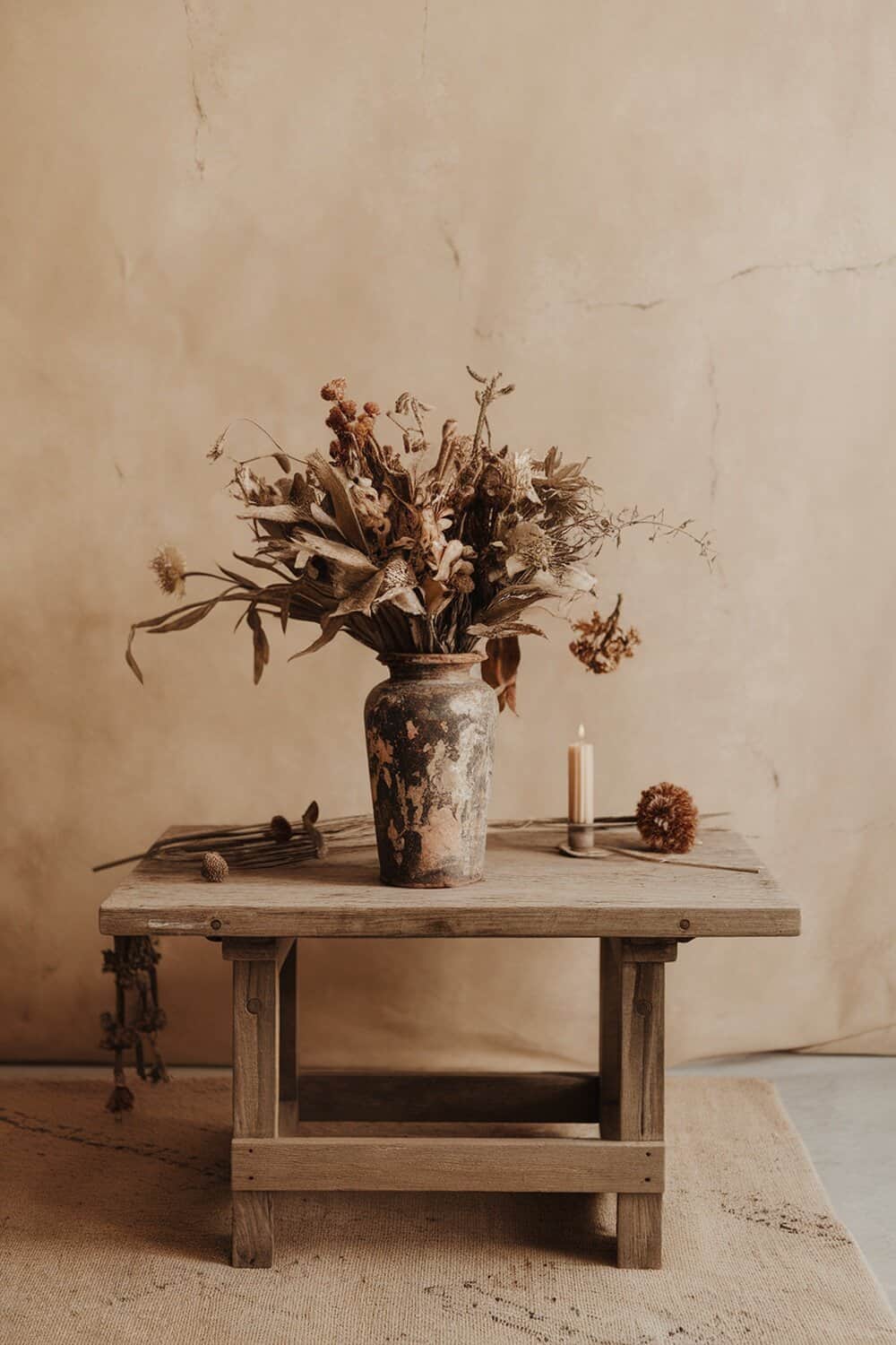 A dried flower bouquet in a rustic vase on a wooden table with a candle.