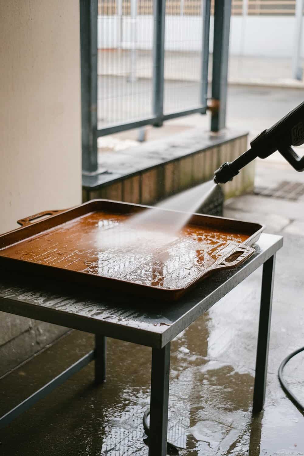 A pressure washer cleaning a dirty cookie sheet on a table.