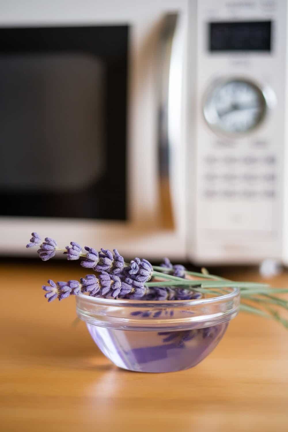 A bowl of lavender water with lavender sprigs in front of a microwave.