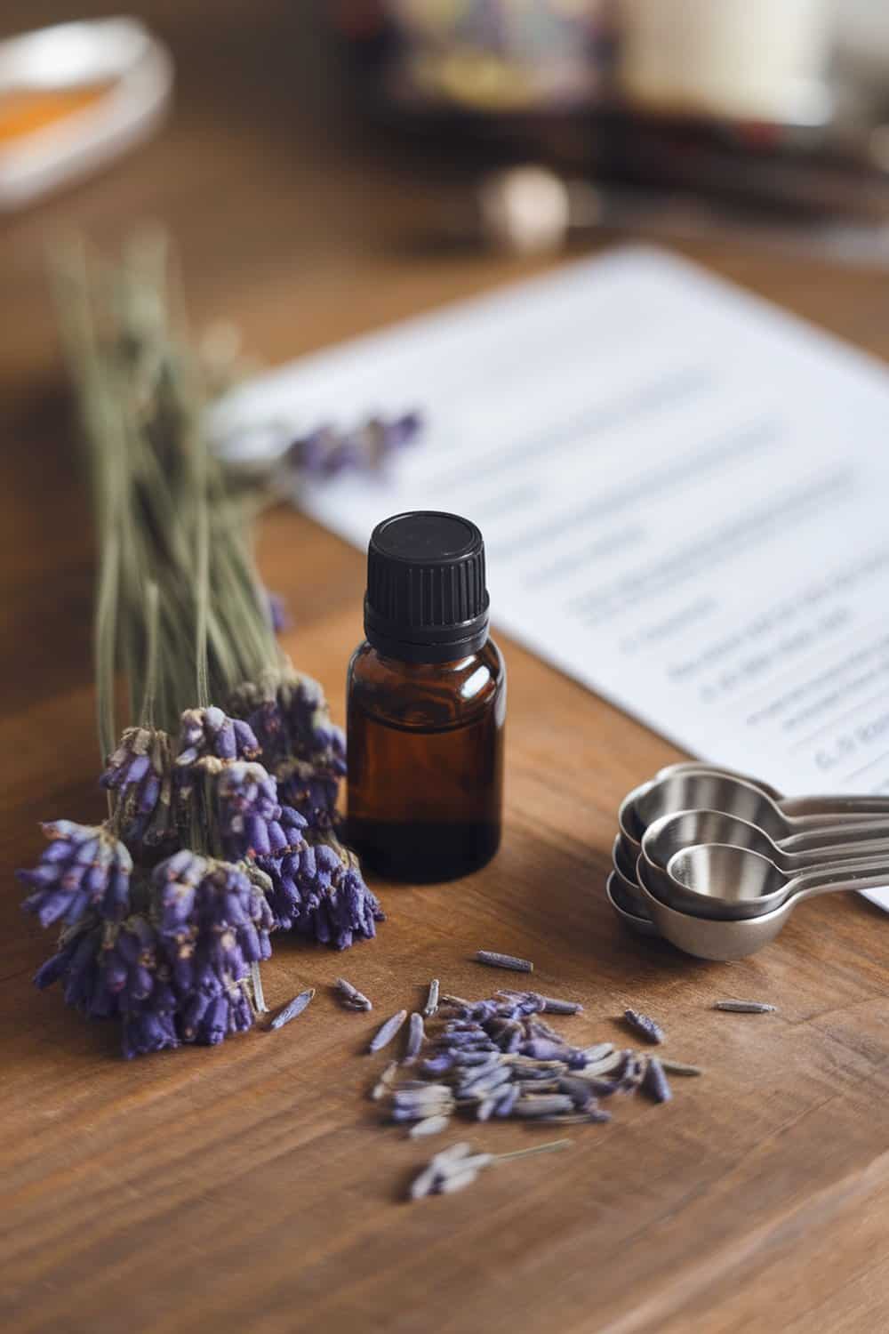 Lavender essential oil bottle with dried lavender flowers and measuring spoons on a wooden table.