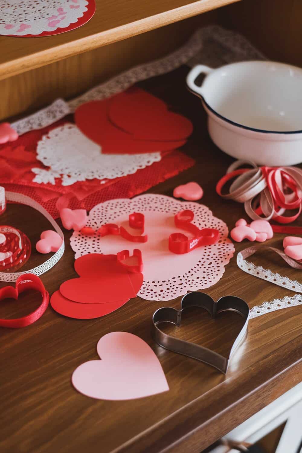 A collection of Valentine's Day craft supplies including heart-shaped cutouts, ribbons, and doilies on a wooden surface.