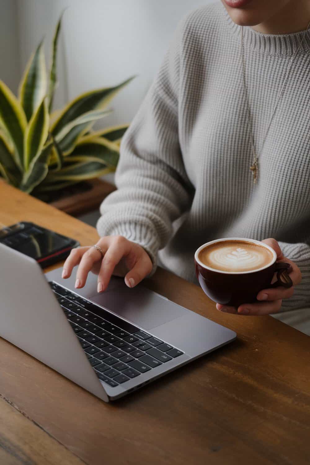 A person typing on a laptop while holding a cup of coffee, with a plant in the background.