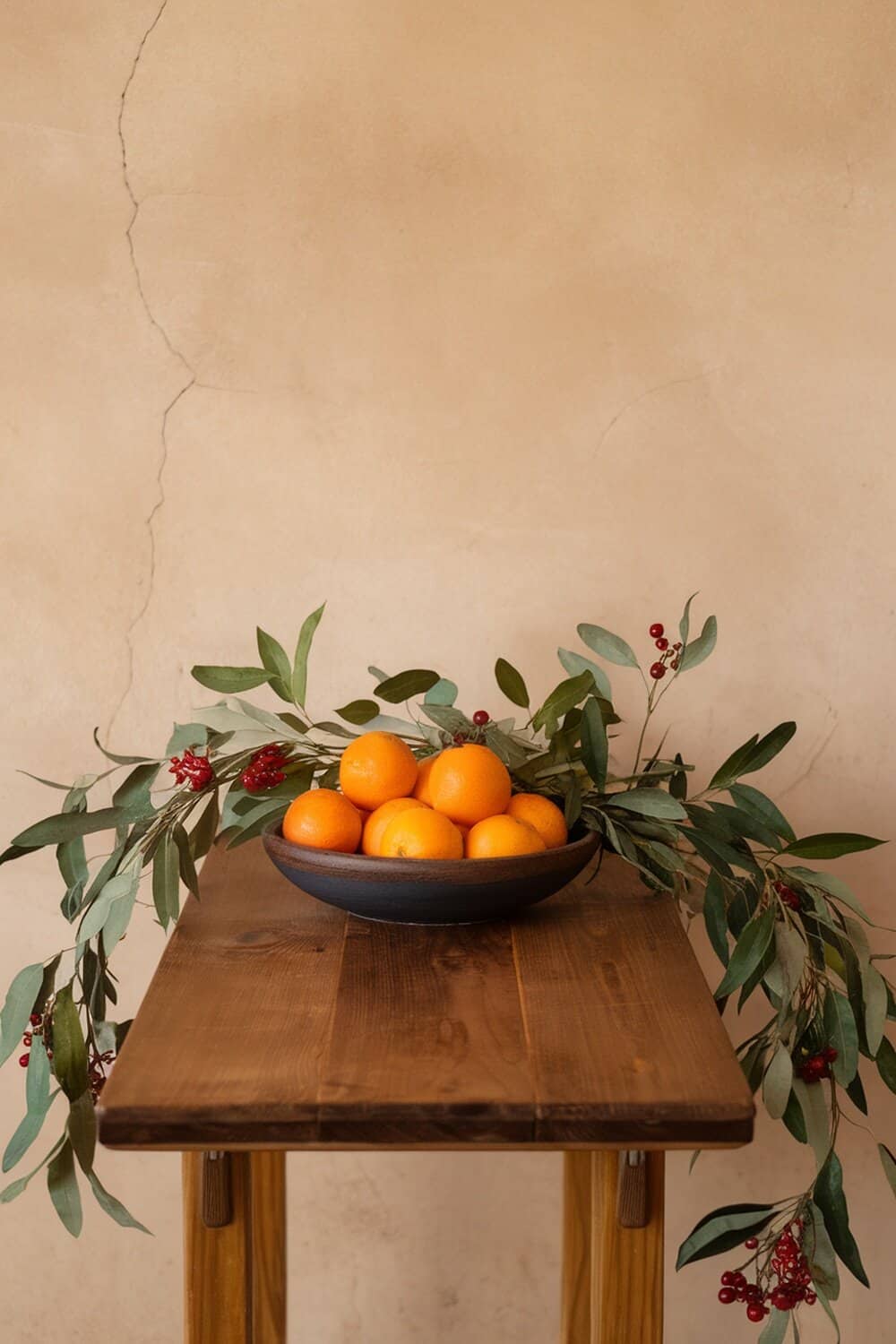 A wooden table with a bowl of oranges surrounded by greenery and red berries.