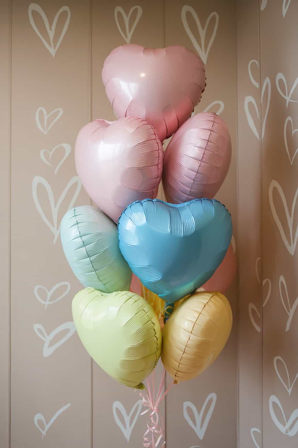 A cluster of colorful heart-shaped balloons in pastel shades against a wall with heart patterns.