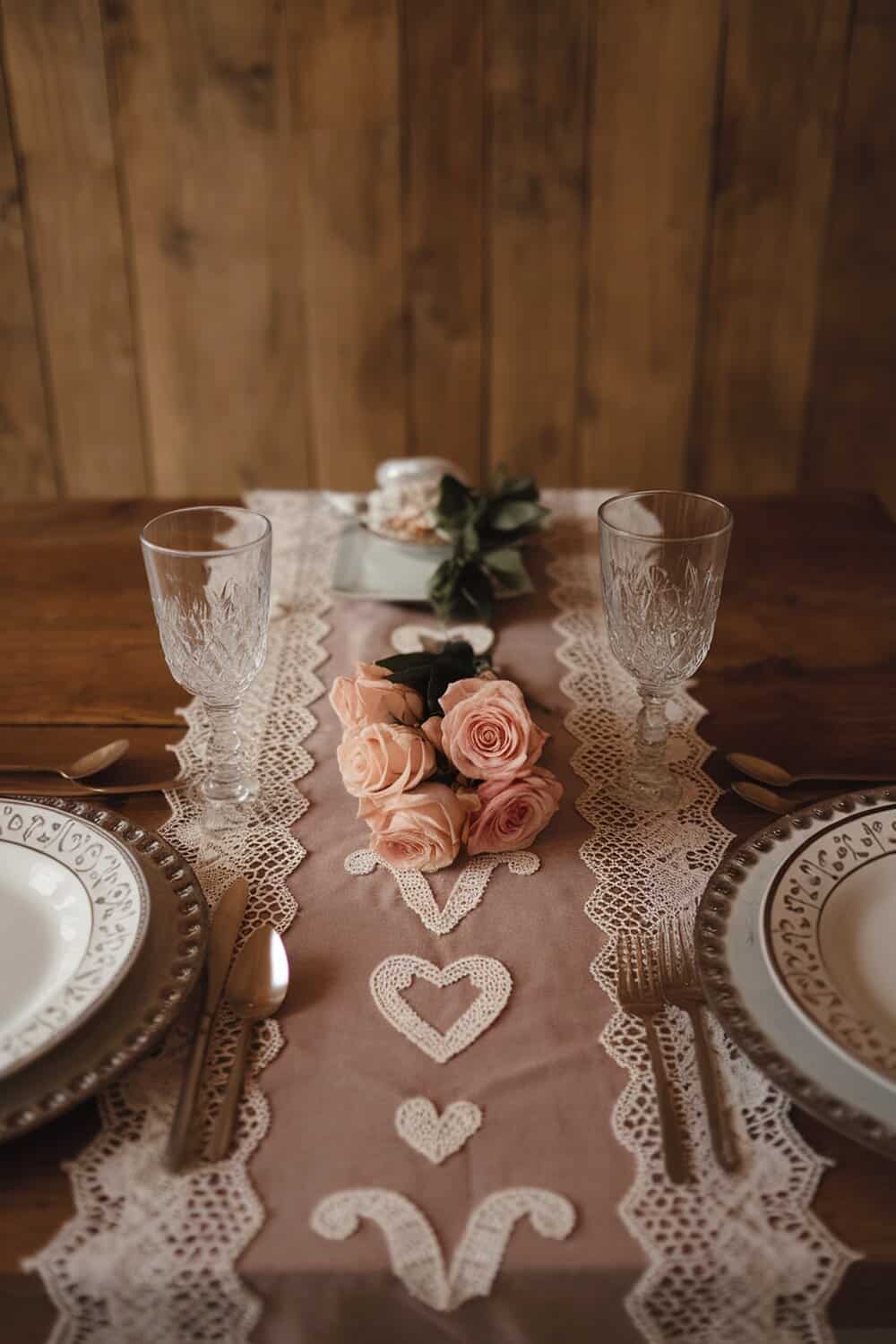 A beautifully set table with a lace table runner, pink roses, and elegant dinnerware.