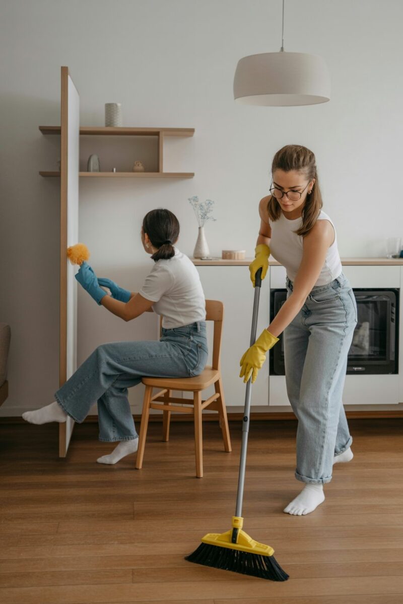 Two friends cleaning a house together, one on a chair dusting and the other sweeping the floor.
